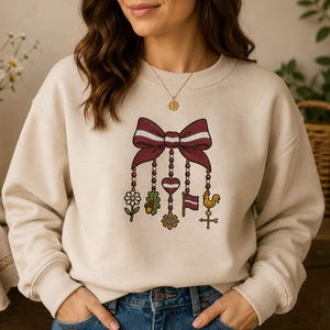 Könnte beinhalten: Ein beigefarbenes Sweatshirt mit Rundhalsausschnitt und einem zentralen Aufdruck einer großen kastanienbraunen Schleife. Von der Schleife hängen fünf Anhänger herab: ein weißes Gänseblümchen, ein Eichenblatt mit Eichel, ein Herz mit kastanienbraunem Streifen, eine kastanienbraune Flagge und eine goldene Wetterfahne in Form eines Hahns.