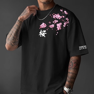 May include: A black short-sleeved t-shirt featuring a graphic of a pink cherry blossom branch extending from the shoulder. The shirt includes the white Japanese kanji character for cherry blossom and the text TOKYO with Japanese characters below it.