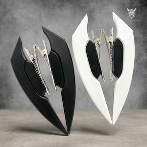 May include: Two hair clips, one black and one white, each with a shield-like design. The clips have a silver metal clasp. The background is a neutral grey.