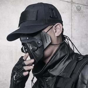 Op de afbeelding: Zwarte tactische pet en gezichtsmasker. Het masker heeft een geometrisch ontwerp met een filter en verstelbare bandjes. De pet heeft een klittenband patch. De afbeelding toont ook een zwarte handschoen en jas. De merknaam "VENTAWEAR" is zichtbaar.