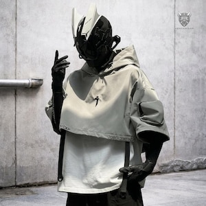 Puede incluir: Una parte superior tipo poncho con capucha de color gris claro con un logotipo negro y el texto "7080/JIYEIND". La parte superior tiene un diseño único con una manga corta y un bolsillo en la manga. El modelo lleva un casco futurista y guantes negros.