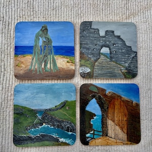 May include: A set of four square coasters featuring hand-painted scenes. One shows a hooded figure by the sea, another a stone archway, the third a green coastal valley with a bay, and the fourth a wooden door set within a stone archway by the ocean.