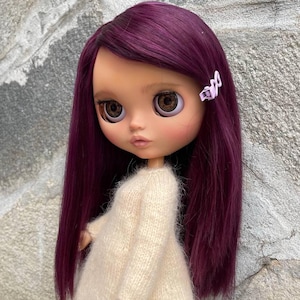 May include: A doll with long, straight, deep purple hair and a light purple hair clip. The doll has large brown eyes, a small nose, and pink lips. It is wearing a long-sleeved, cream-coloured, fuzzy knit sweater that covers the torso and arms.