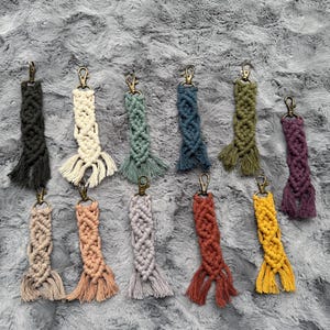 May include: A collection of macrame keychains in various colors, including black, cream, green, blue, olive, purple, tan, peach, gray, rust, and yellow. Each keychain features a woven design and a gold-colored clasp.