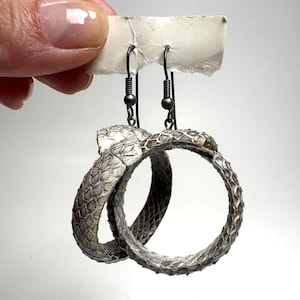 May include: A pair of hoop earrings featuring a textured, grey and white snakeskin pattern. The circular hoops are suspended from dark metal fishhook-style ear wires, creating a continuous loop design that mimics the appearance of a coiled snake.