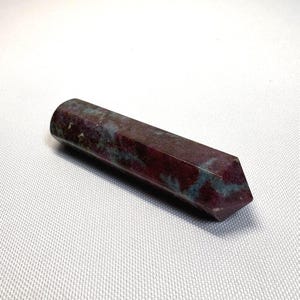May include: A polished, elongated gemstone with a faceted end. The stone features a deep burgundy color with streaks of teal and a cylindrical shape. The gemstone is approximately 4 inches long.
