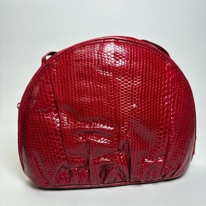 May include: A semi-circular handbag in a vibrant red colour with a glossy, textured finish that mimics a snakeskin pattern. The bag features a structured top zip closure and subtle vertical pleating along the bottom edge of the front panel.