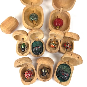 May include: A collection of small, hinged wooden boxes, each containing a hand-painted miniature figurine. The figures include various beetles, ladybugs, and turtles in vibrant colours like red, green, and black. One box is closed, showing a textured exterior.
