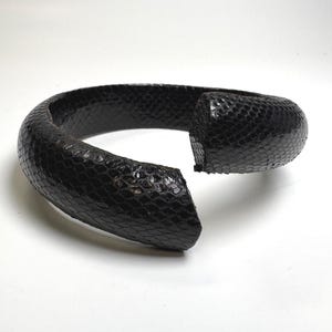 May include: A thick, open-ended cuff bracelet featuring a dark black, textured snakeskin pattern. The bracelet has a rounded, substantial profile with a glossy finish and a distinct gap between the two ends, showcasing the detailed scale texture.