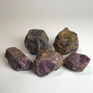 May include: Five rough ruby gemstones with a mix of dark purple, brown, and gold hues. The raw stones have a natural, unpolished appearance, showcasing their unique textures and colors. These gemstones are ideal for jewelry making or collecting.