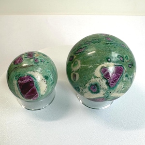 Peut inclure: Deux sphères polies en pierres précieuses. La plus grande sphère mesure environ 7,6 cm de diamètre, et la plus petite environ 5 cm. Les deux présentent une couleur de base verte avec des inclusions rouges et blanches, reposant sur des socles en acrylique transparent.