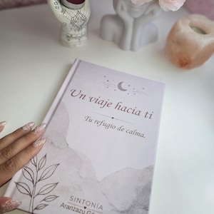 Può includere: Un libro con copertina rigida caratterizzato da una copertina color lavanda chiaro e bianco ad acquerello, con un'illustrazione a linee di una foglia, una luna crescente e il testo "Un viaje hacia ti, Tu refugio de calma, SINTONÍA Aranzazu Gómez" in grigio scuro.