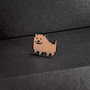 May include: A pixelated dog enamel pin with a tan body and black outline. The pin is a square shape with a dog silhouette. The pin is on a dark gray surface.