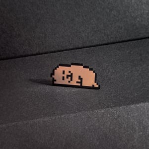 May include: A pixelated pin featuring a light brown figure in a resting pose, outlined in black. The pin is set against a dark gray, textured background, creating a contrast that highlights the design.