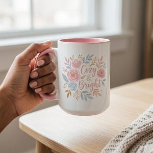 Może przedstawiać: Biały ceramiczny kubek z różowym wnętrzem i uchwytem. Kubek jest ozdobiony kwiatowymi wzorami w kolorze różowym, niebieskim i złotym oraz napisem "Cozy & Bright". Kubek jest trzymany przez osobę.