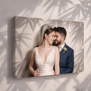 Puede incluir: Impresión en lienzo de una pareja, posiblemente una foto de boda. La mujer lleva un vestido blanco y una tiara, el hombre un traje azul. La imagen está montada en una pared, con sombras de hojas como fondo.