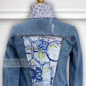 May include: A denim jacket with a floral patterned back panel and collar. The back panel features a light blue background with a floral design in shades of blue, green, and yellow. The collar has a blue and white floral pattern. The text "ONE OF A KIND Salvaged Threads" is visible.