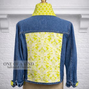 May include: A blue denim jacket with a vibrant yellow and white patterned back panel and collar. The cuffs are also lined with the same yellow pattern. The text "ONE OF A KIND Salvaged Threads" is printed on the lower left side.