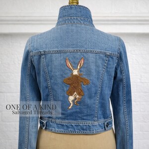 May include: A light blue denim jacket with a high collar and button closures. The back features a detailed embroidered rabbit wearing a brown coat. The text "ONE OF A KIND Salvaged Threads" is visible on the lower left.