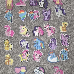 Peut inclure: Une collection de breloques de chaussures My Little Pony colorées. Les breloques présentent divers personnages de poneys dans différentes poses et couleurs, notamment rose, violet, bleu et jaune. Certaines breloques ont des accents arc-en-ciel et l'inscription "My Little Pony".