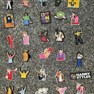 Könnte beinhalten: Eine Sammlung bunter Schuhanhänger mit verschiedenen Designs. Die Anhänger zeigen Bilder eines Hauses, einer Kiwi und Figuren in verschiedenen Outfits. Einige Anhänger tragen den Text "HARRY'S HOUSE" und "LOVE ON TOUR".