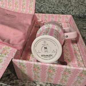 May include: A pink floral Stanley mug with a matching handle and lid, presented in a decorative box with a pink floral pattern. The mug is designed in Seattle, WA, and holds 1.18L. The box also contains a pink satin bag.