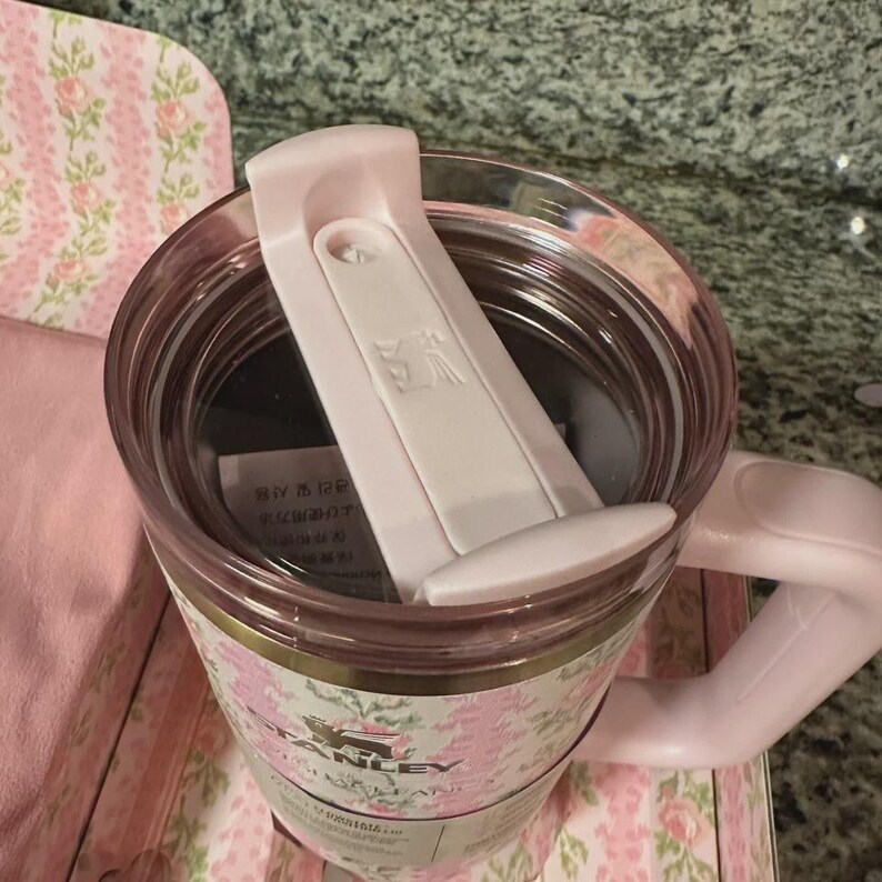 May include: A pink floral Stanley travel mug with a white lid. The mug has a handle and a clear interior. The floral pattern includes pink roses and green leaves. The mug is sitting on a pink floral patterned surface.