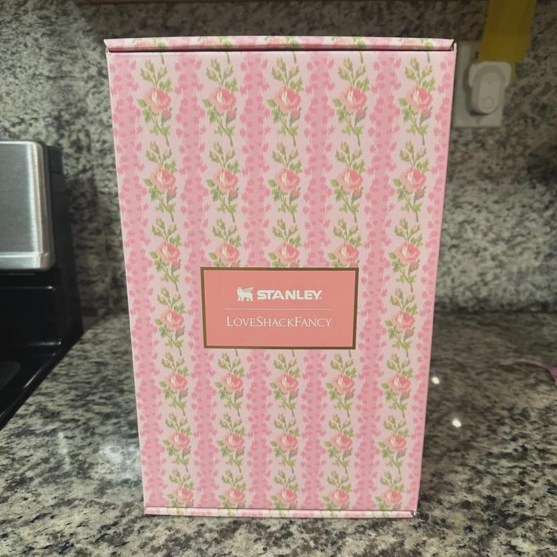 May include: A rectangular box with a pink floral pattern. The box features a repeating design of pink roses and green leaves on a light pink background. The text "STANLEY" and "LOVESHACKFANCY" are displayed on a gold label.