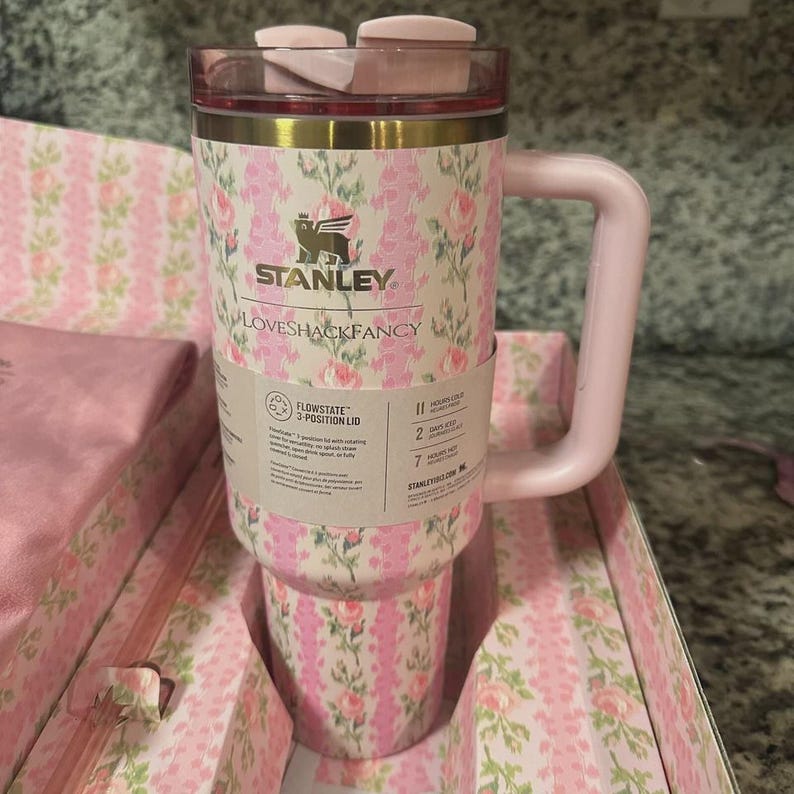 May include: A pink and floral Stanley Loveshackfancy travel mug with a handle. The mug has a pink lid and a floral pattern with the Stanley logo. The mug is in a floral patterned box.