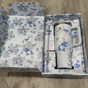 Puede incluir: Un vaso Stanley gris claro con asa, adornado con un estampado floral azul y blanco, se presenta en una caja con estampado floral a juego. El vaso tiene un borde dorado y la marca es visible. La caja y el vaso est&aacute;n sobre una superficie de madera.