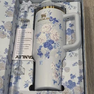 Puede incluir: Un vaso Stanley LoveShackFancy azul claro con asa, adornado con un estampado floral en blanco y azul. El vaso est&aacute; en su caja original, que tambi&eacute;n presenta el mismo dise&ntilde;o floral. Incluye una pajita.