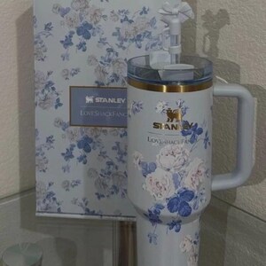 Puede incluir: Un vaso Stanley azul claro y su caja a juego, ambos adornados con un estampado floral en blanco y azul. El vaso tiene un asa y una tapa transparente. El texto "STANLEY LOVESHACKFANCY" es visible.
