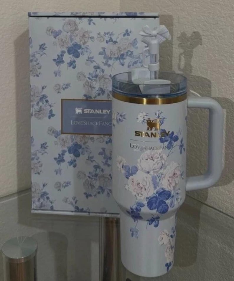 Puede incluir: Un vaso Stanley azul claro y su caja a juego, ambos adornados con un estampado floral en blanco y azul. El vaso tiene un asa y una tapa transparente. El texto "STANLEY LOVESHACKFANCY" es visible.