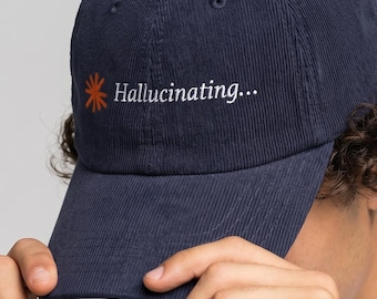 May include: Navy blue corduroy baseball cap with the text "Hallucinating..." in white script and a stylized orange sunburst graphic. The cap has a curved brim and a button on top.