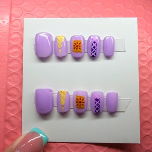 May include: A set of ten press-on nails in a light purple shade, arranged in two rows on a white card. The designs include a yellow braided hair pattern, a yellow and orange sun lantern motif, and a purple crisscross corset-style lace pattern.