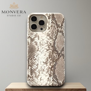 Pode incluir: Capa de smartphone com padrão de pele de cobra em tons de castanho e creme. A capa tem um recorte para a câmara e é exibida num bloco de madeira. A marca "MONVERA STUDIO CO" é visível no canto superior esquerdo.