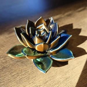 May include: A three-dimensional succulent sculpture featuring layered, pointed petals with a metallic finish that shifts between deep navy blue, emerald green, and bronze gold tones, creating a vibrant, multi-colored appearance.