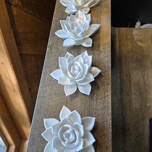 May include: Four white, multi-layered lotus flower ornaments arranged in a vertical line. Each flower features pointed, overlapping petals with a smooth, matte finish. The ornaments are identical in shape and color, displaying a symmetrical design.