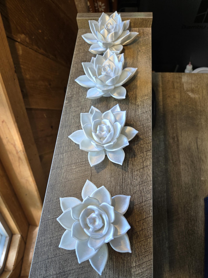 May include: Four white, multi-layered lotus flower ornaments arranged in a vertical line. Each flower features pointed, overlapping petals with a smooth, matte finish. The ornaments are identical in shape and color, displaying a symmetrical design.
