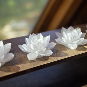 May include: Four white, translucent, carved stone lotus flower sculptures arranged in a row. Each flower features multiple layered, pointed petals radiating from a central point, creating a symmetrical, blooming appearance with a smooth, polished finish.