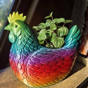 May include: A chicken-shaped planter featuring a vibrant rainbow gradient finish that transitions from yellow at the comb to green, blue, purple, and red across the body. The hollow center holds a small green plant with heart-shaped leaves.