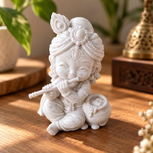 May include: A white speckled figurine of a child deity wearing an ornate turban with a peacock feather, playing a flute. The figure is seated in a cross-legged position next to a small pot filled with round objects, featuring detailed facial expressions.