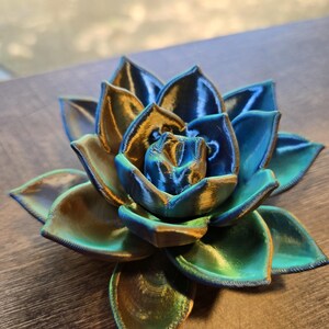 May include: A 3D printed succulent sculpture featuring layered, pointed petals in a metallic gradient of deep blue, teal, and gold. The object displays a distinct ribbed texture from the printing process, with a symmetrical, blooming floral shape.