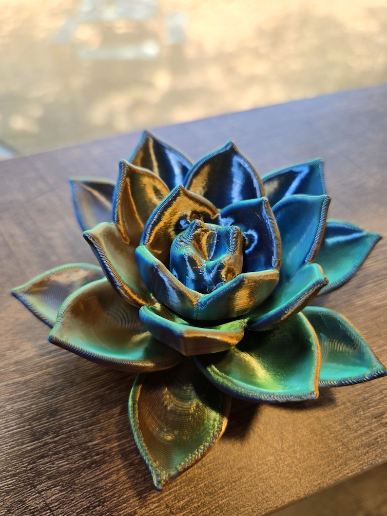 May include: A 3D printed succulent sculpture featuring layered, pointed petals in a metallic gradient of deep blue, teal, and gold. The object displays a distinct ribbed texture from the printing process, with a symmetrical, blooming floral shape.