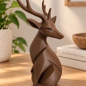 May include: A brown deer figurine with a smooth, angular surface and geometric lines. The deer is depicted in a seated position with its head turned to the side, featuring prominent antlers and closed eyes, crafted with a matte finish.