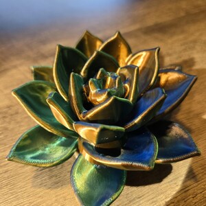 May include: A 3D printed succulent sculpture featuring layered, pointed petals with a metallic finish that shifts between deep blue, vibrant green, and warm gold tones. The object has a visible textured surface from the printing process.
