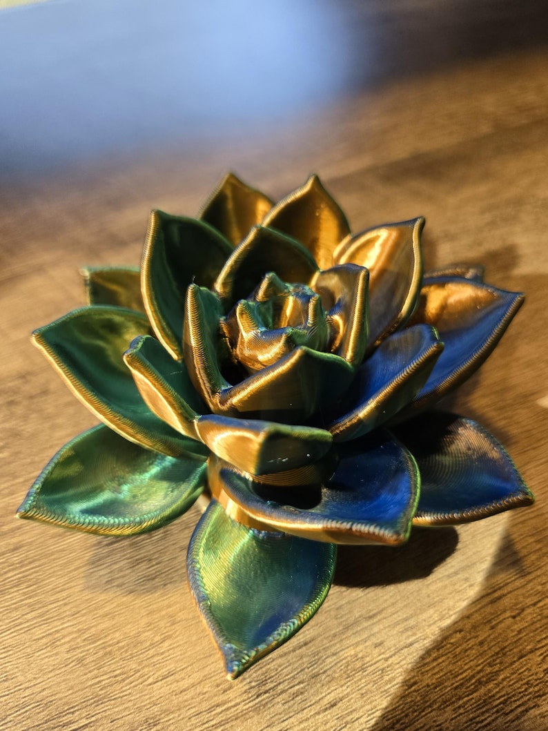 May include: A 3D printed succulent sculpture featuring layered, pointed petals with a metallic finish that shifts between deep blue, vibrant green, and warm gold tones. The object has a visible textured surface from the printing process.