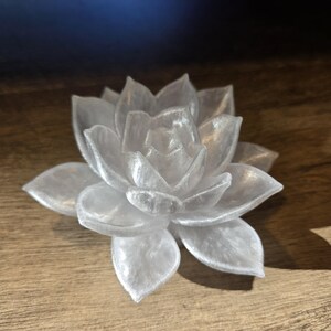 May include: A translucent white succulent figurine with a pearlescent finish. The object is shaped like a blooming flower with multiple layered, pointed petals radiating from a central point, creating a realistic botanical appearance.
