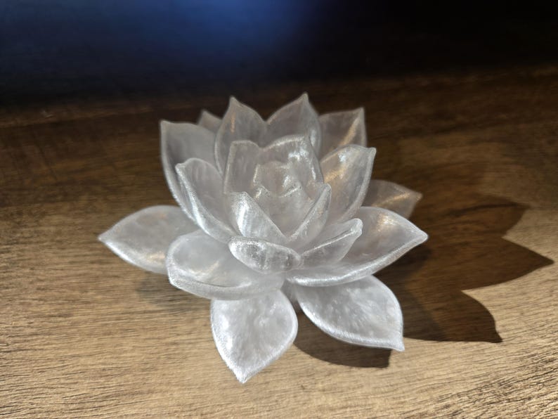 May include: A translucent white succulent figurine with a pearlescent finish. The object is shaped like a blooming flower with multiple layered, pointed petals radiating from a central point, creating a realistic botanical appearance.