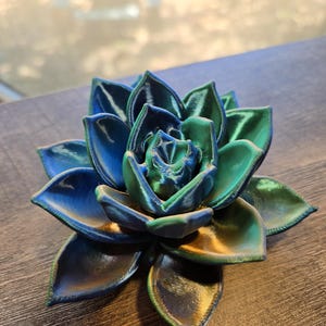 May include: A 3D printed succulent sculpture featuring layered, pointed petals in a gradient of iridescent blue, green, and gold tones. The object has a visible textured surface from the printing process, with a symmetrical, blooming rosette shape.